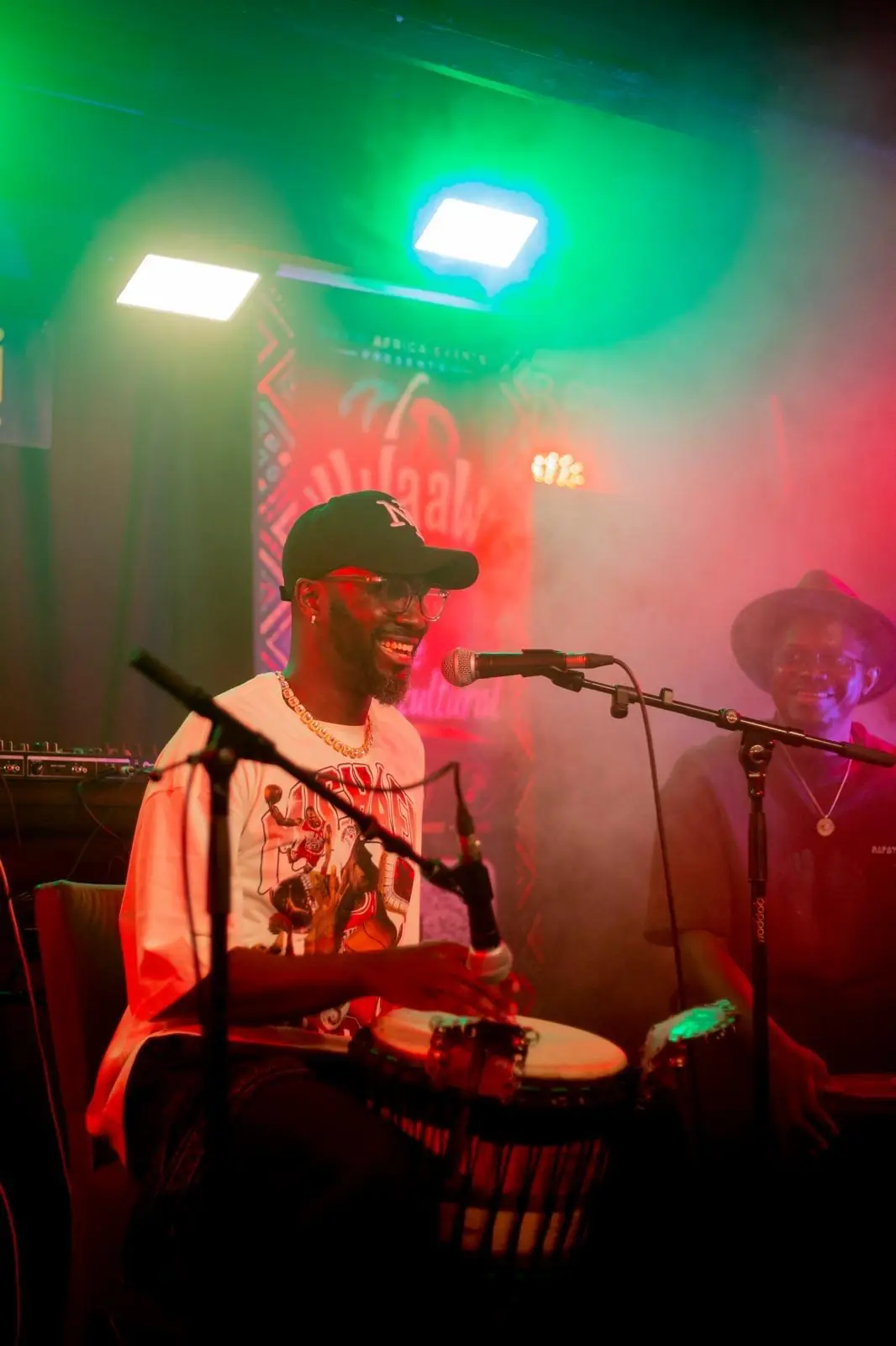 A drummer plays djembe on stage under green and red lights