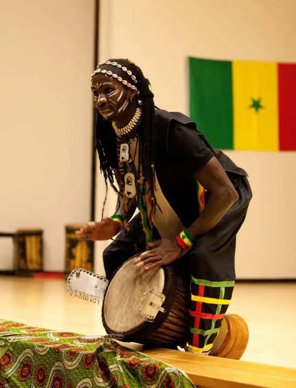 A drummer with painted face plays djembe in front of the Senegalese flag