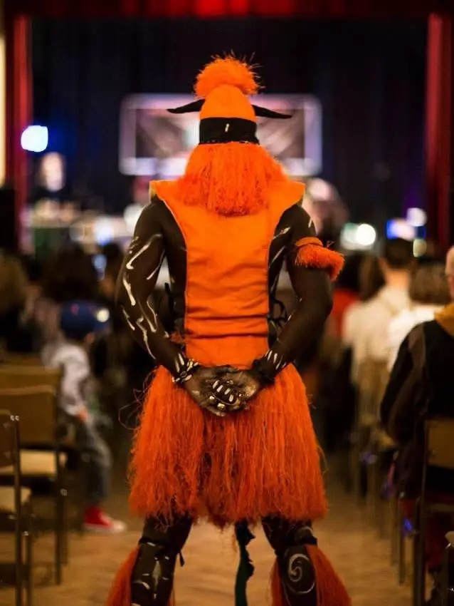 A masked dancer in a vivid orange costume faces the audience