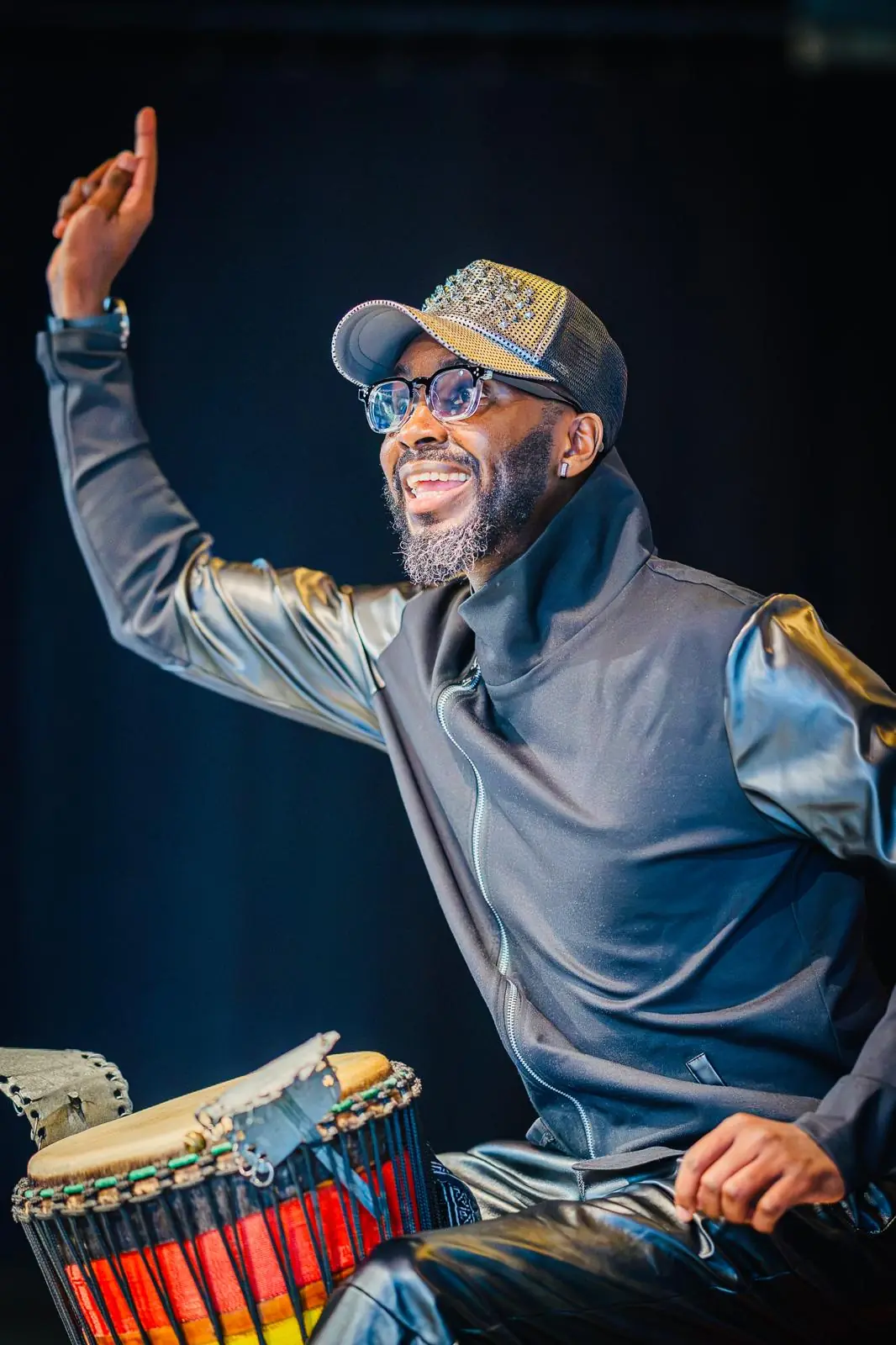 A smiling percussionist raises his hand mid-performance beside a colourful djembe