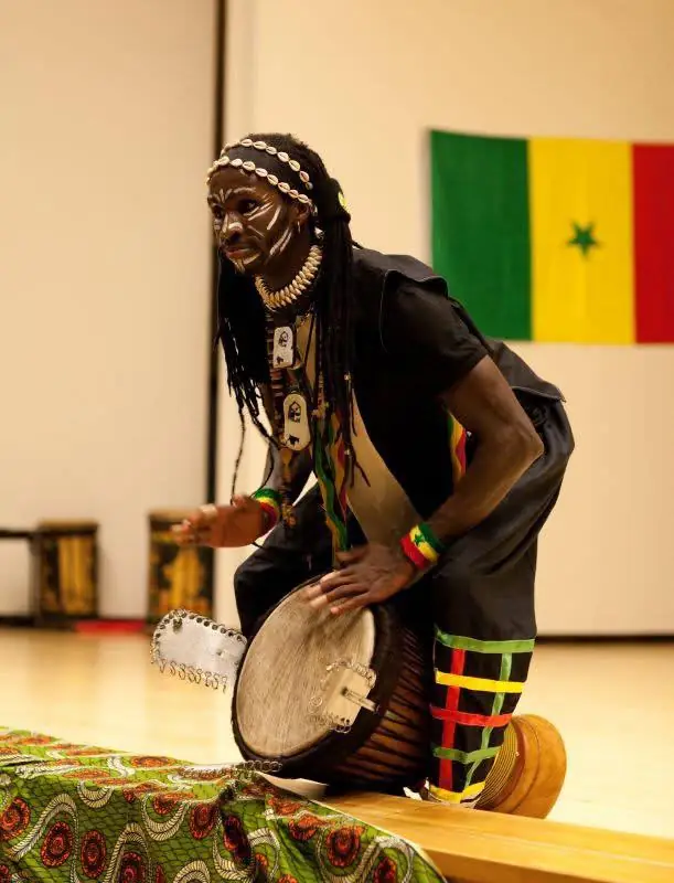 A drummer with painted face plays djembe in front of the Senegalese flag